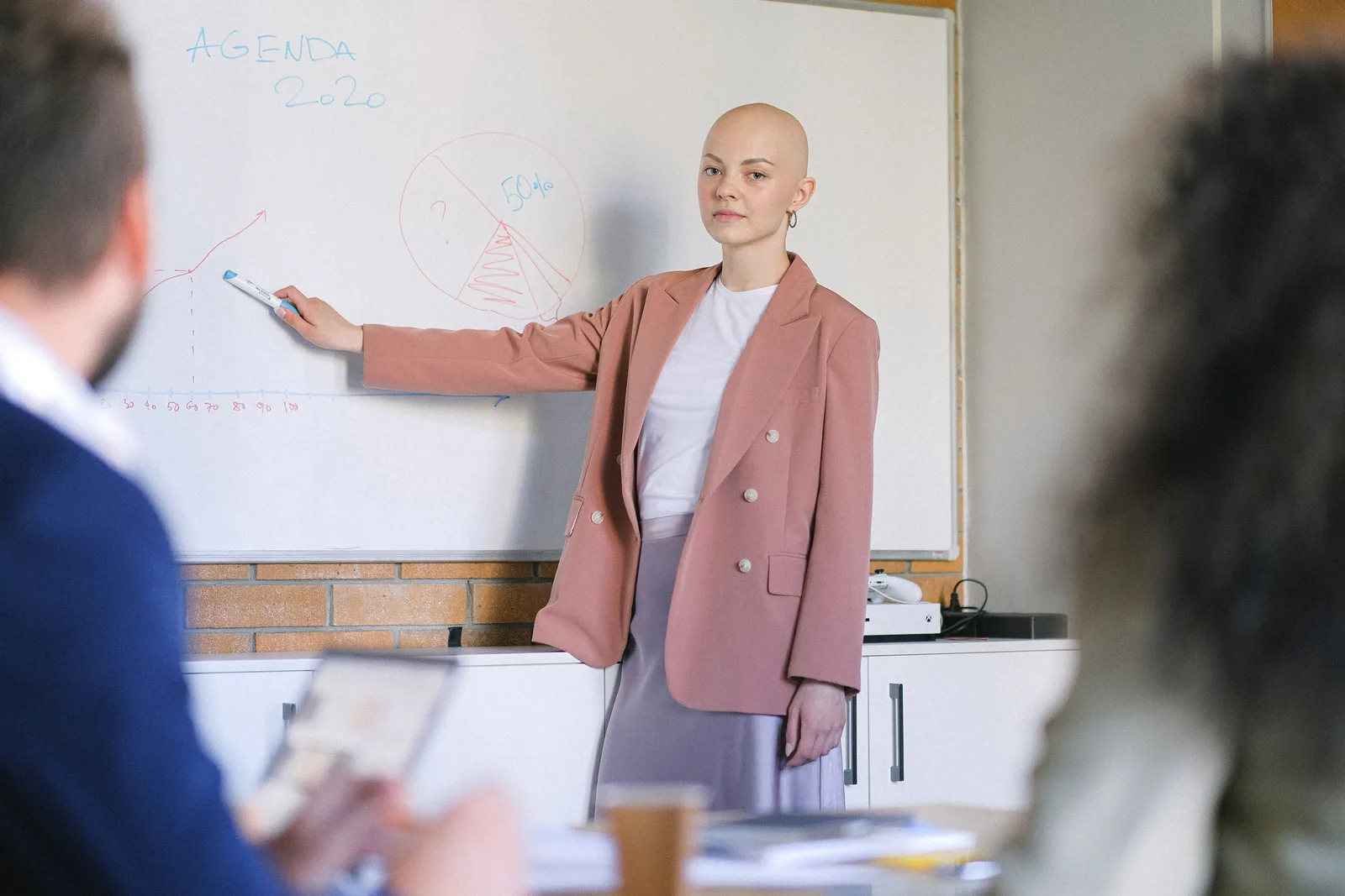 Content female looking at camera and pointing with marker at whiteboard with diagrams while explaining marketing plan to colleagues in conference room during briefing